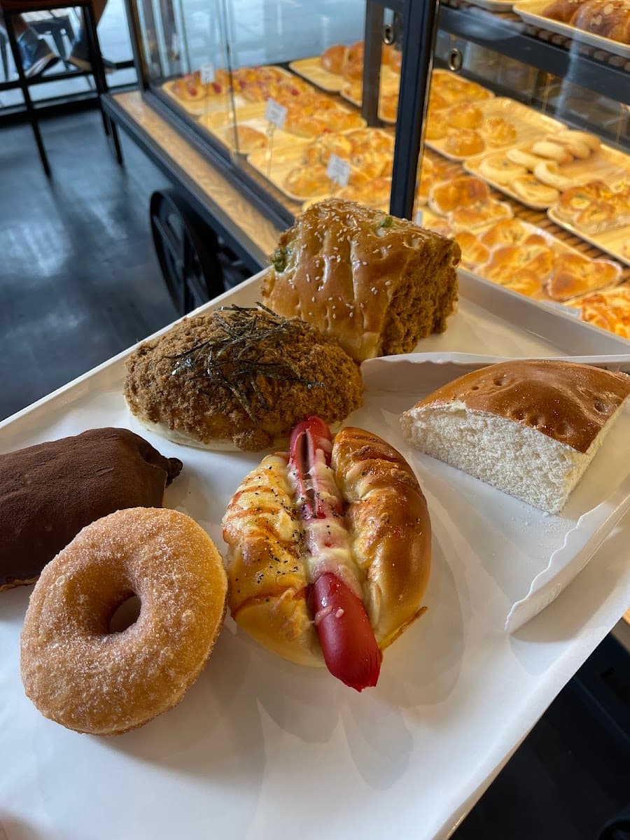 A white bakery tray holds an assortment of pastries, including a sugar doughnut, hot dog bun, and savory pork floss bread in front of a display case.